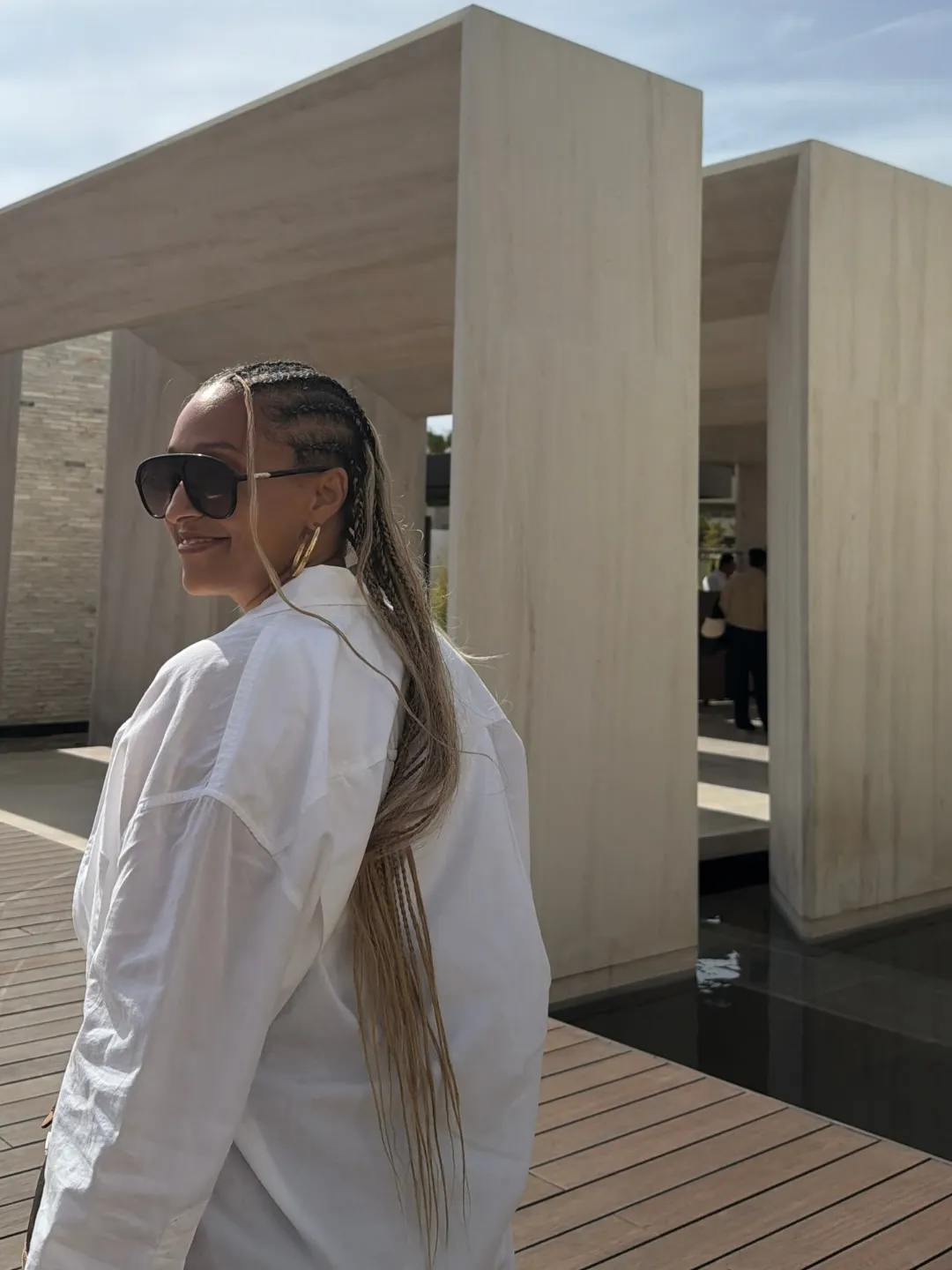 Tia Mowry smiling in sunglasses with long braids, wearing a white shirt, on a wooden pathway surrounded by concrete structures.