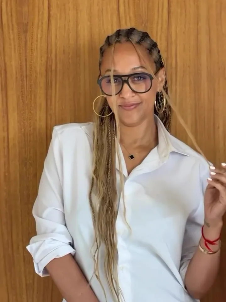 Tia Mowry wearing a white shirt and glasses, smiling at the camera.