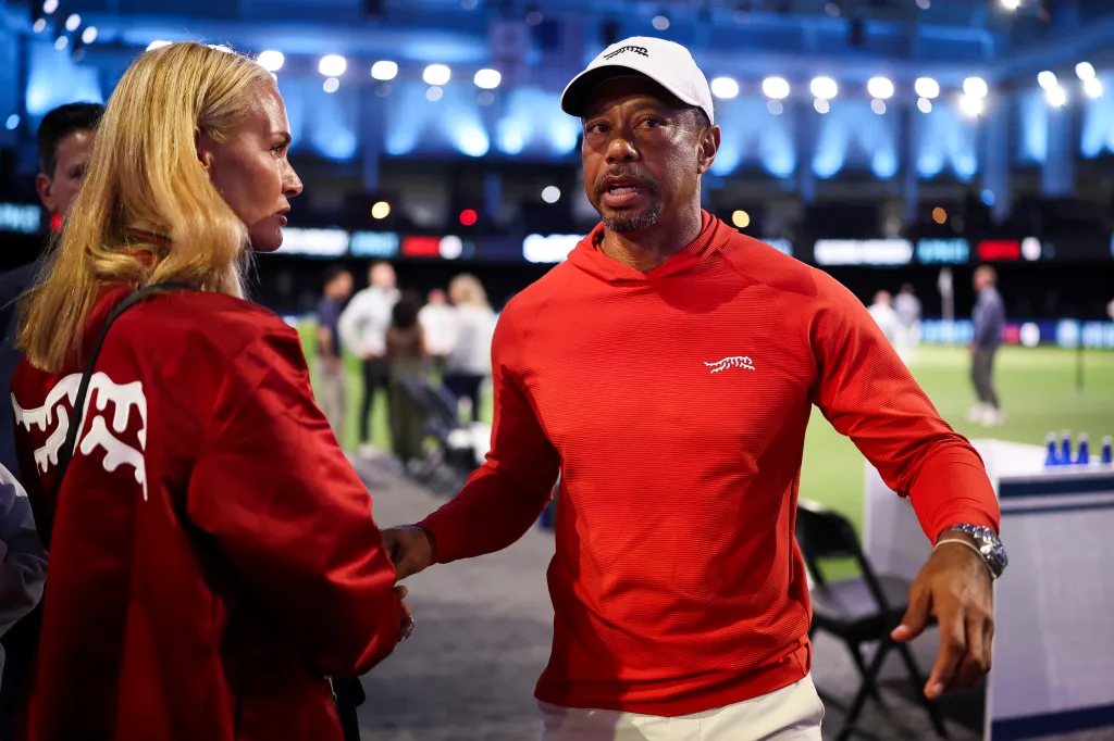 Tiger Woods, wearing a red long-sleeved shirt and white cap, speaks to his girlfriend, Vanessa Trump, who wears a red jacket.
