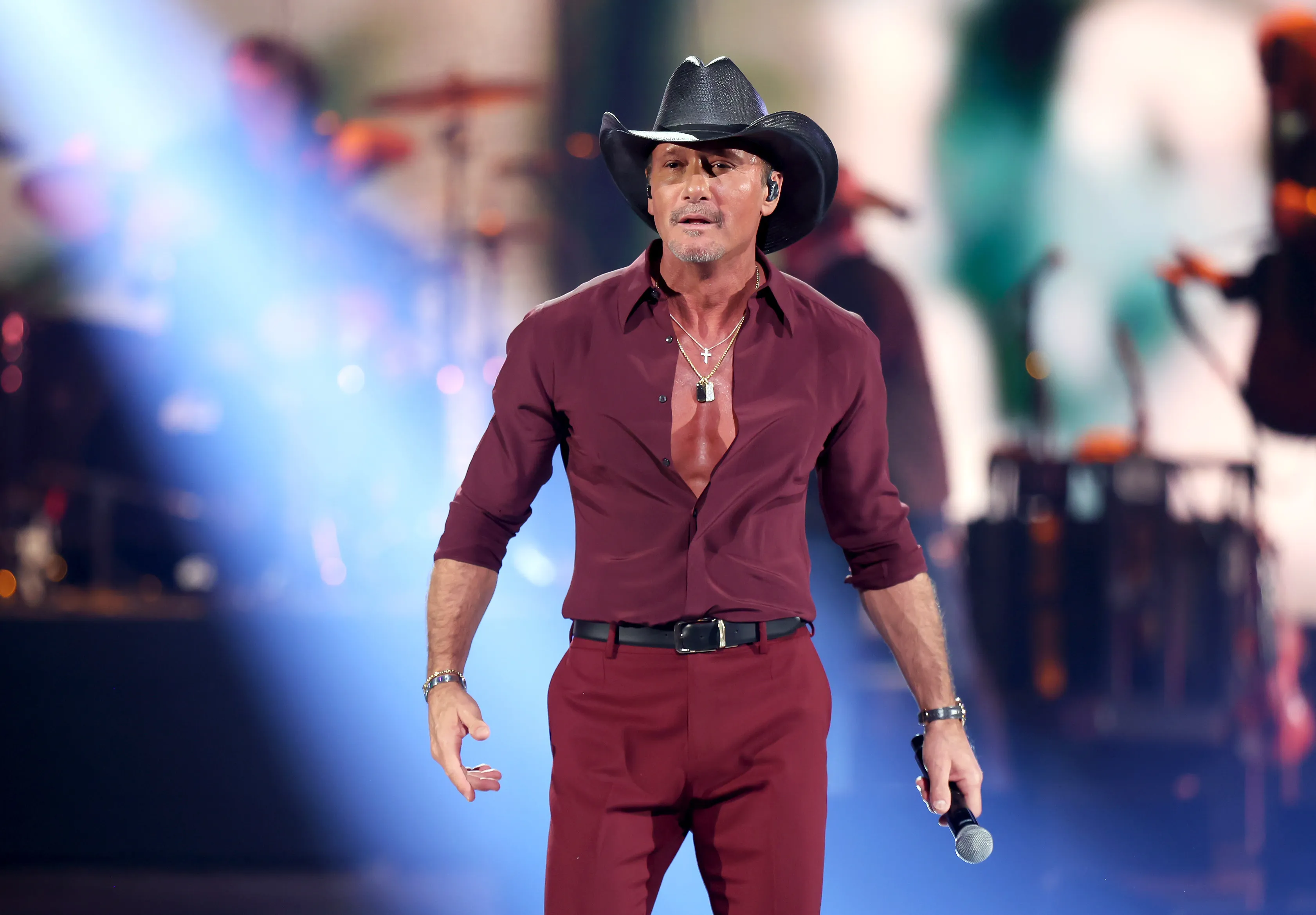 Tim McGraw performing onstage in a maroon shirt, matching pants, and a black cowboy hat, holding a microphone.