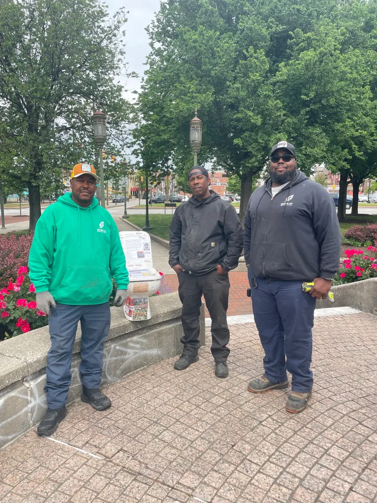 Three men stand beside a toilet decorated with historical text, replacing a Christopher Columbus statue.