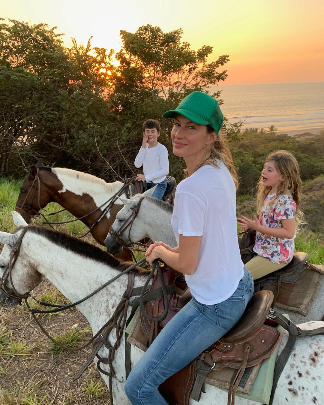 Gisele Bündchen, wearing a green cap, rides a horse with her son and daughter at sunset, overlooking a beach.