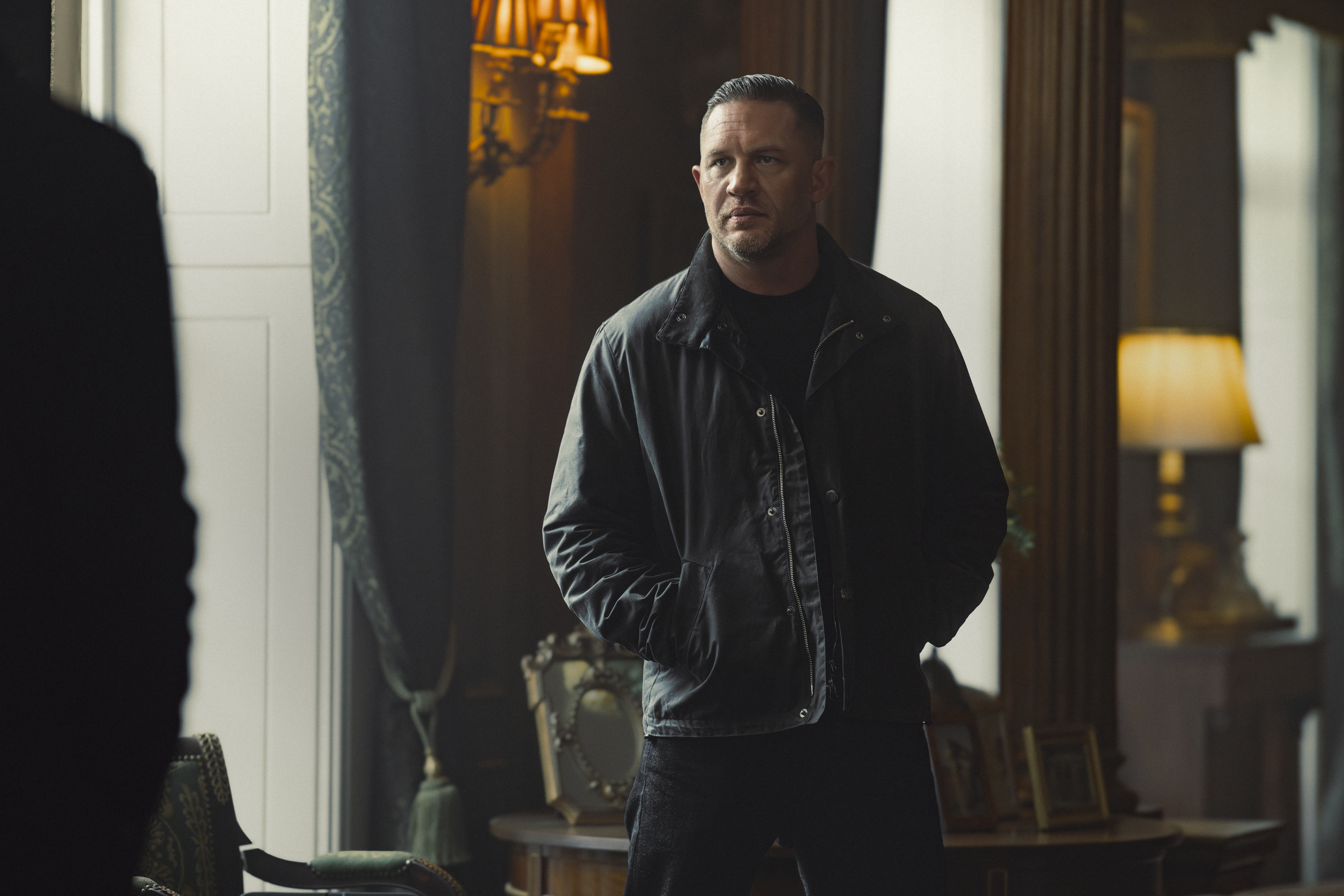 Tom Hardy as Harry Da Souza looking serious in a dark room with ornate furniture and drapes.