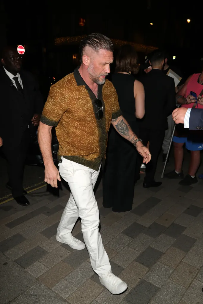 Tom Hardy in a patterned bronze short-sleeved shirt, white trousers, and white sneakers, with a dark tattoo on his left forearm, looking downcast at an afterparty.