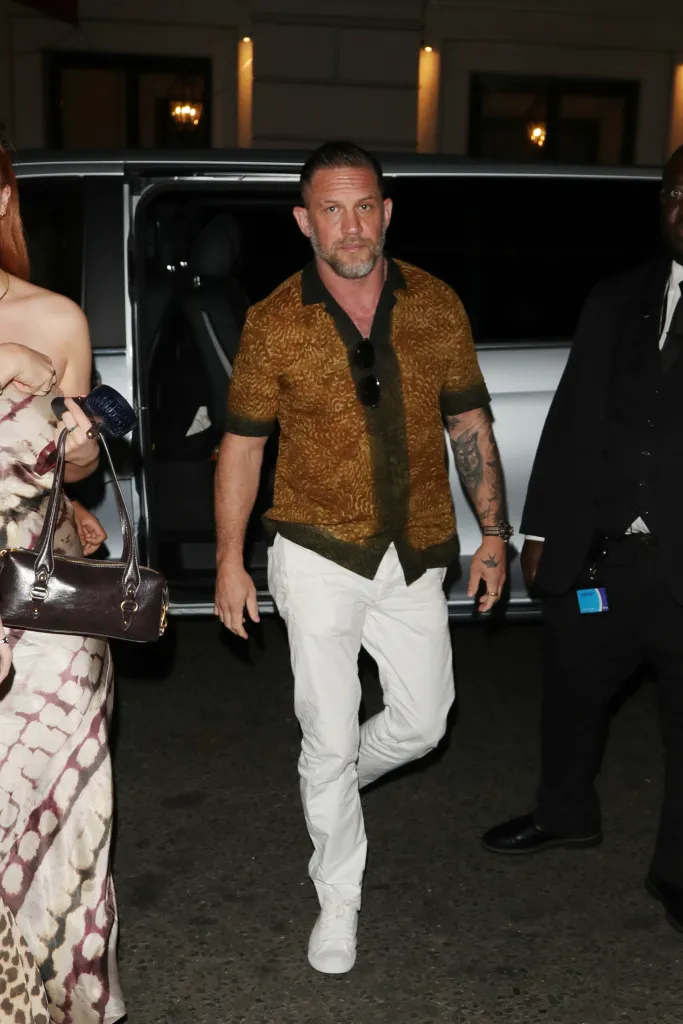 Tom Hardy in a patterned Dries Van Noten shirt, white trousers, and white sneakers, with a woman beside him, walking in London.
