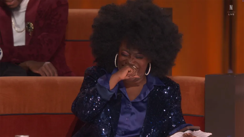 Sheryl Underwood, in a sequined blue jacket, covers her mouth, looking distressed, with her eyes closed and brows furrowed.