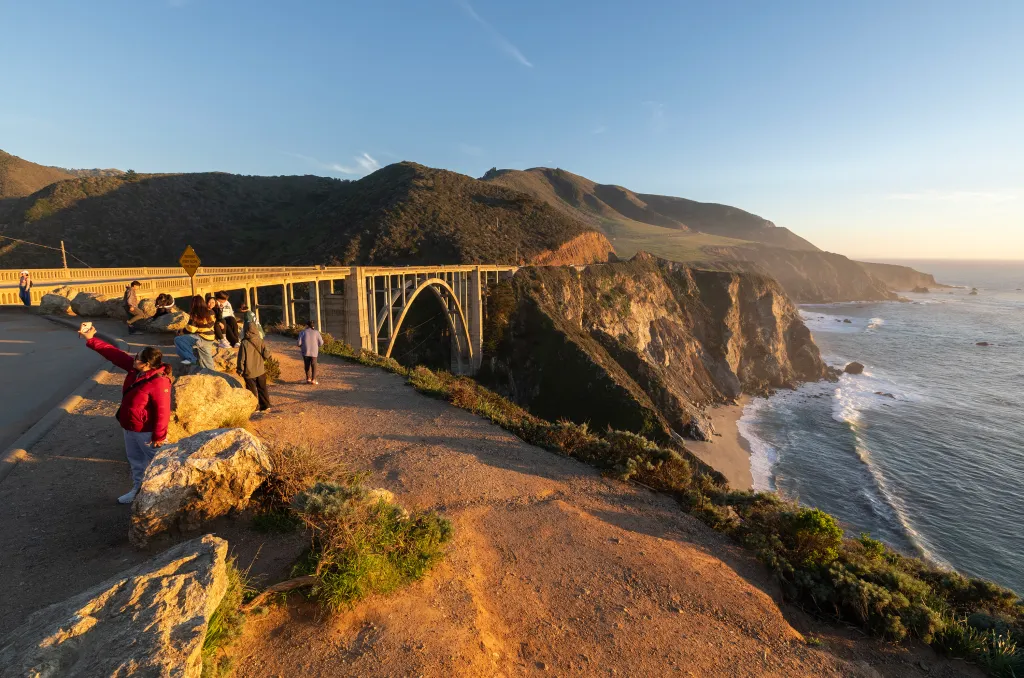 Tourists at Bixby Bridge overlook a cliff, with ocean waves and a beach visible.