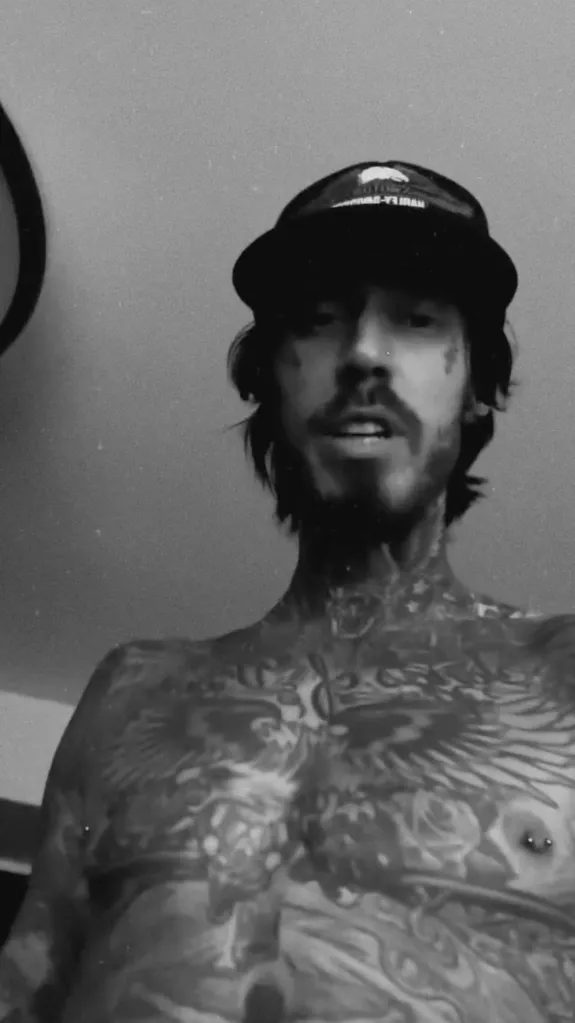 Trace Cyrus, a man with a beard and tattoos across his chest and neck, wearing a baseball cap.