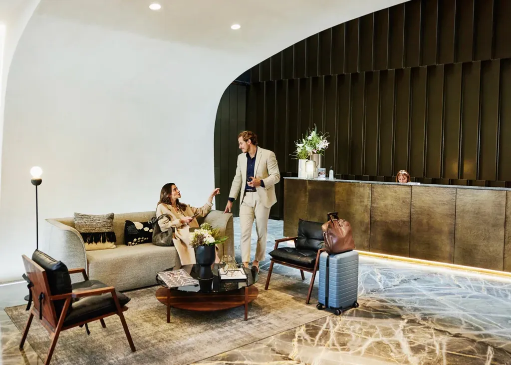 A man and woman talking in a modern hotel lobby, with a suitcase nearby.