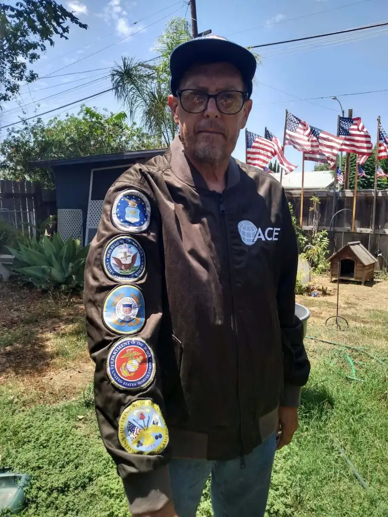 Kerry Sheron wearing a jacket with military patches and American flags in the background.