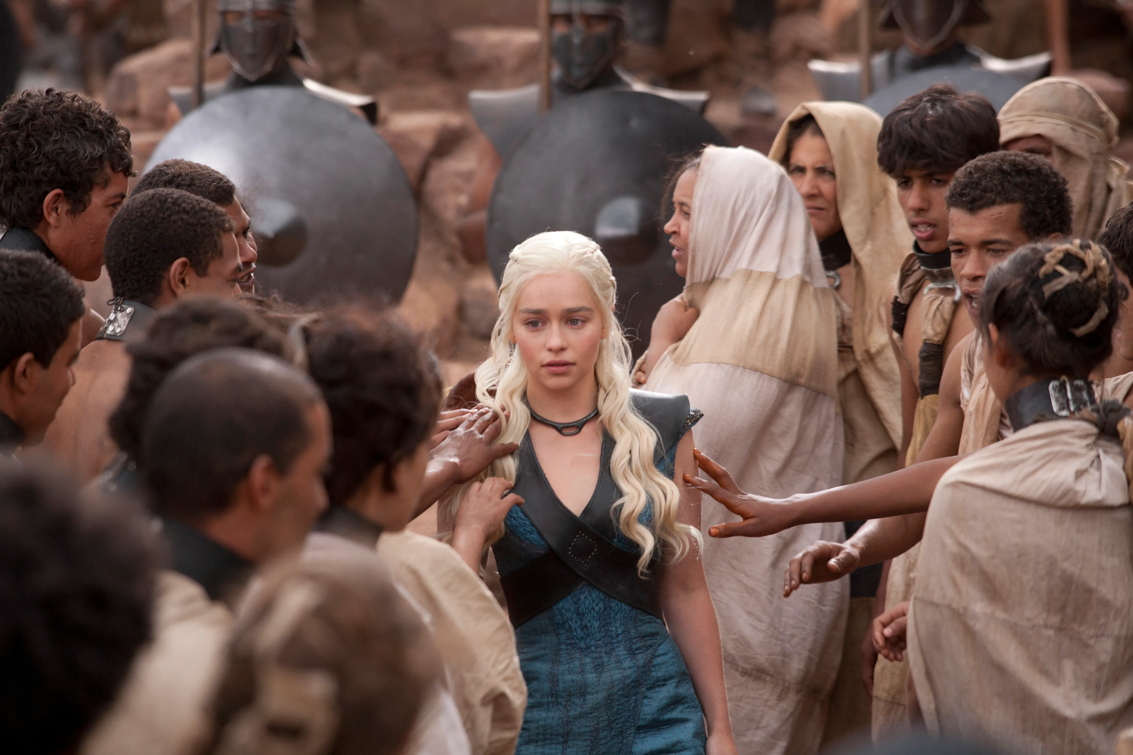 Emilia Clarke as Daenerys Targaryen stands among a crowd, wearing a blue dress and black top.