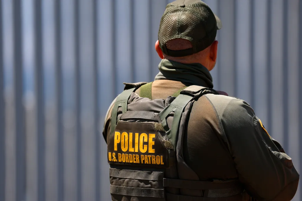 A U.S. Border Patrol agent stands with their back to the camera, wearing a tactical vest with 