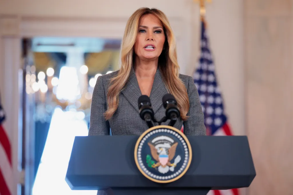 Melania Trump speaking at a podium with two microphones and the Presidential Seal.