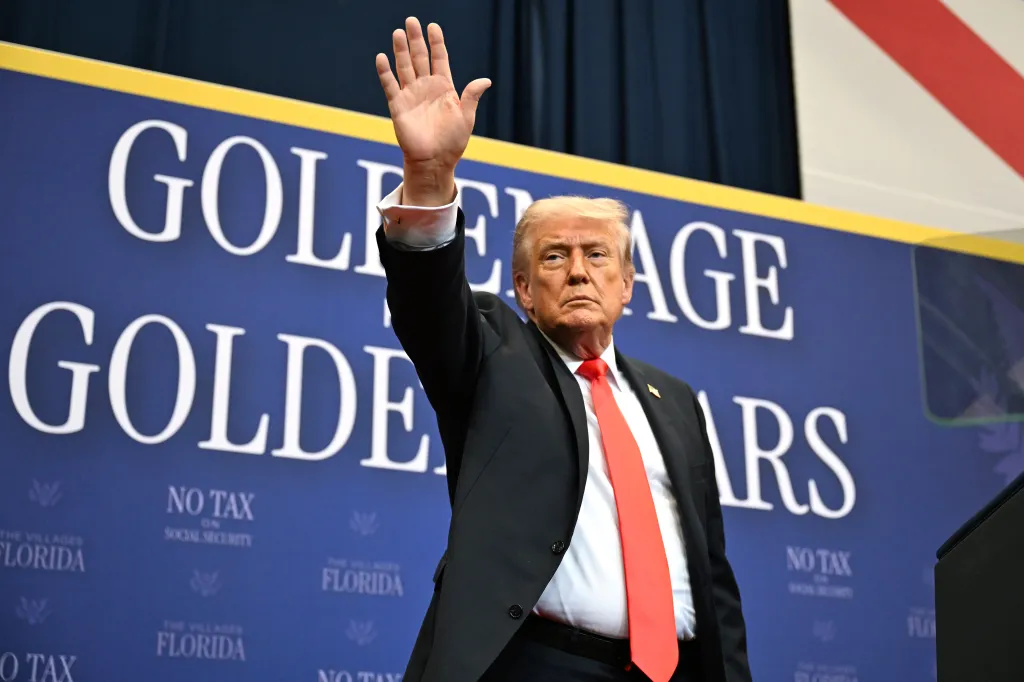 President Trump raises his hand while standing in front of a blue banner that reads 