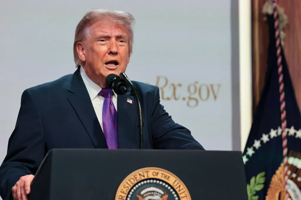 President Donald Trump speaking at a podium with a microphone, with a screen displaying 