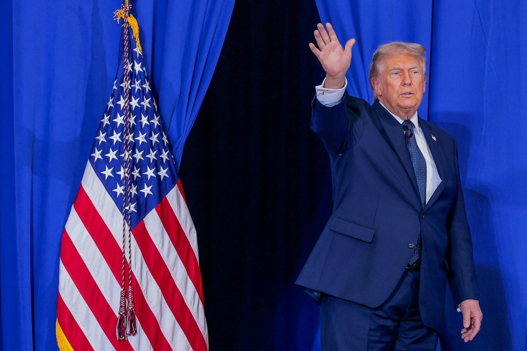 President Donald Trump waves while standing next to an American flag.
