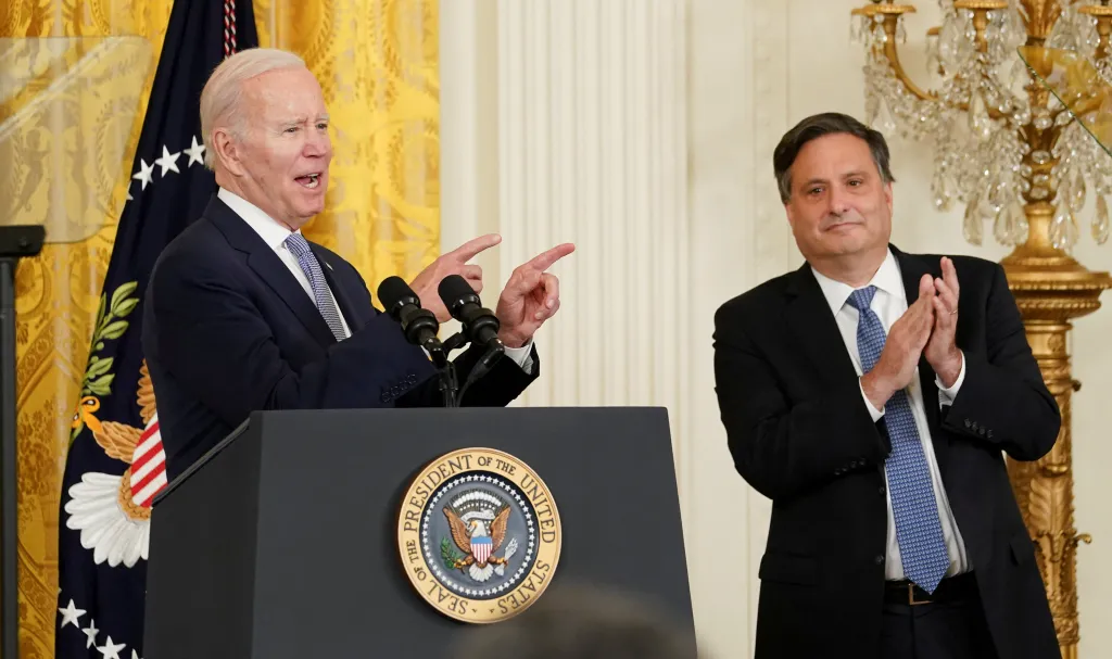 U.S. President Joe Biden speaking at a podium, gesturing with both hands, while Ron Klain stands next to him, clapping.