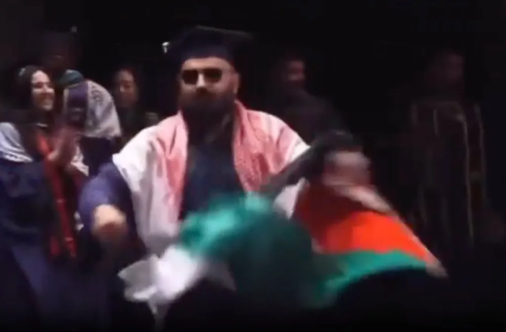 A graduate in cap and gown, wearing sunglasses and a keffiyeh, holding a Palestinian flag.