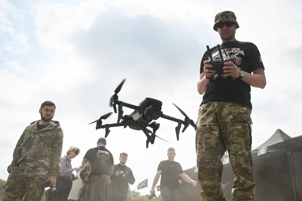 A Ukrainian serviceman in a bucket hat and camouflage pants pilots a drone with a remote control, surrounded by other people and a tent.