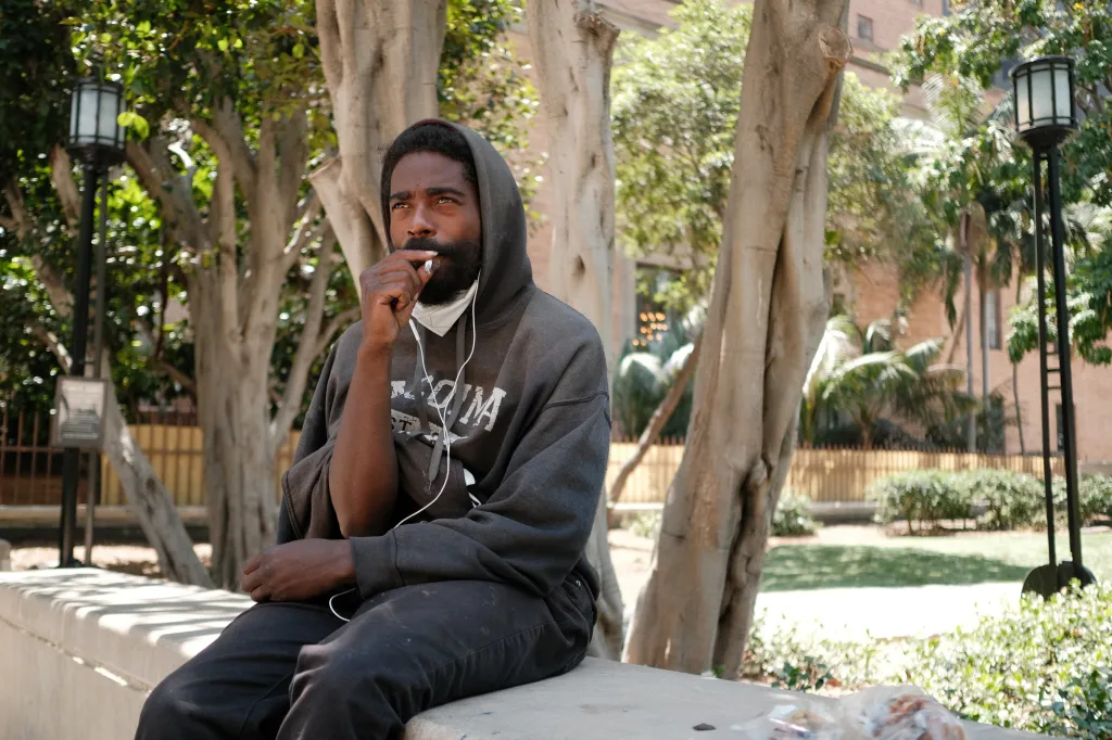 A man with a beard and wearing a gray hoodie with a mask around his neck, sits outdoors with a cigarette in his mouth, looking up thoughtfully.