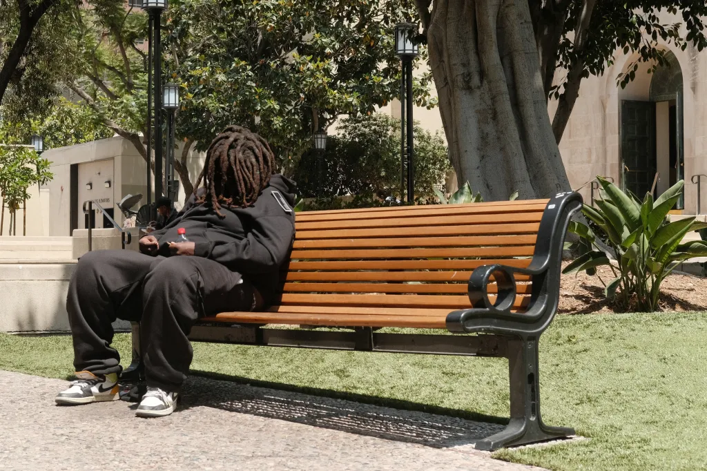 A person with dreadlocks, wearing a black hoodie and sweatpants, sits on a wooden bench outside the Los Angeles Central Library.
