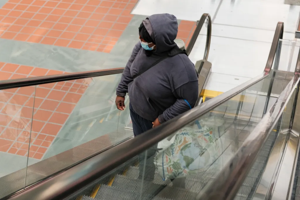 A person wearing a mask and a hooded sweatshirt riding an escalator with a reusable bag.