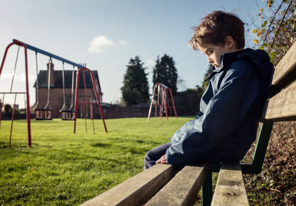 A lonely boy sitting on a park bench with his head down, facing an empty playground.