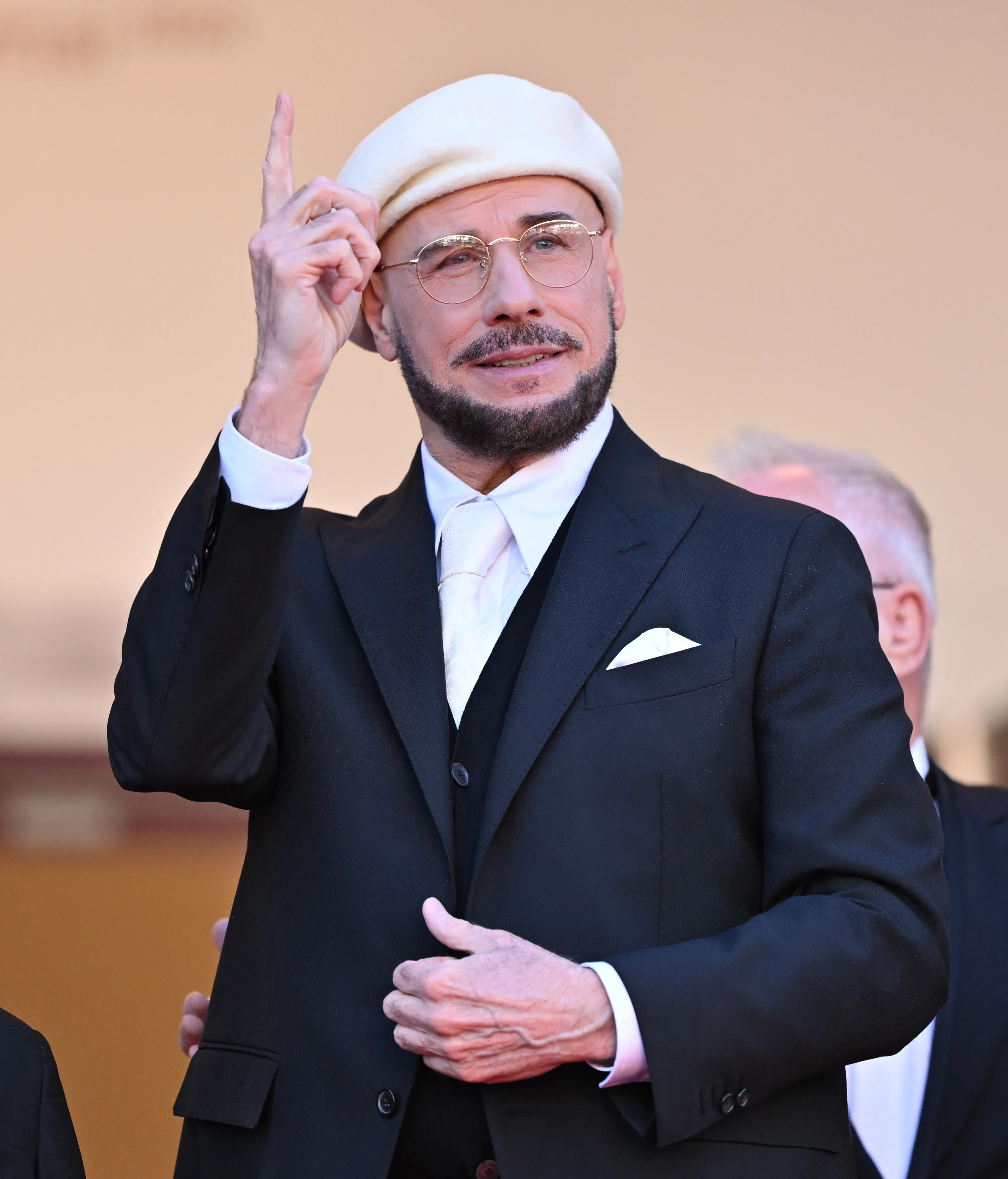 Actor John Travolta in a white beret, light pink tie, and black suit, pointing upwards at the 79th Cannes Film Festival.