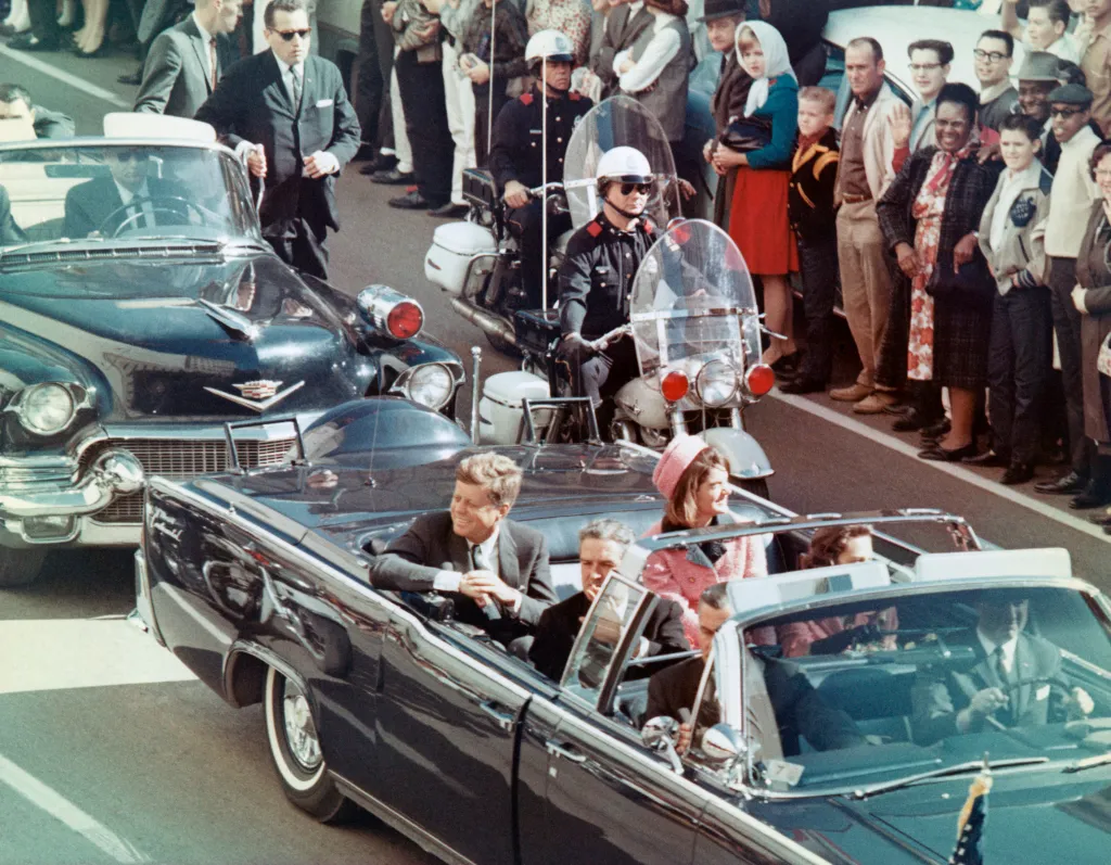 President John F. Kennedy, First Lady Jacqueline Kennedy, and Governor John Connally wave to crowds from a convertible car during a motorcade in Dallas.