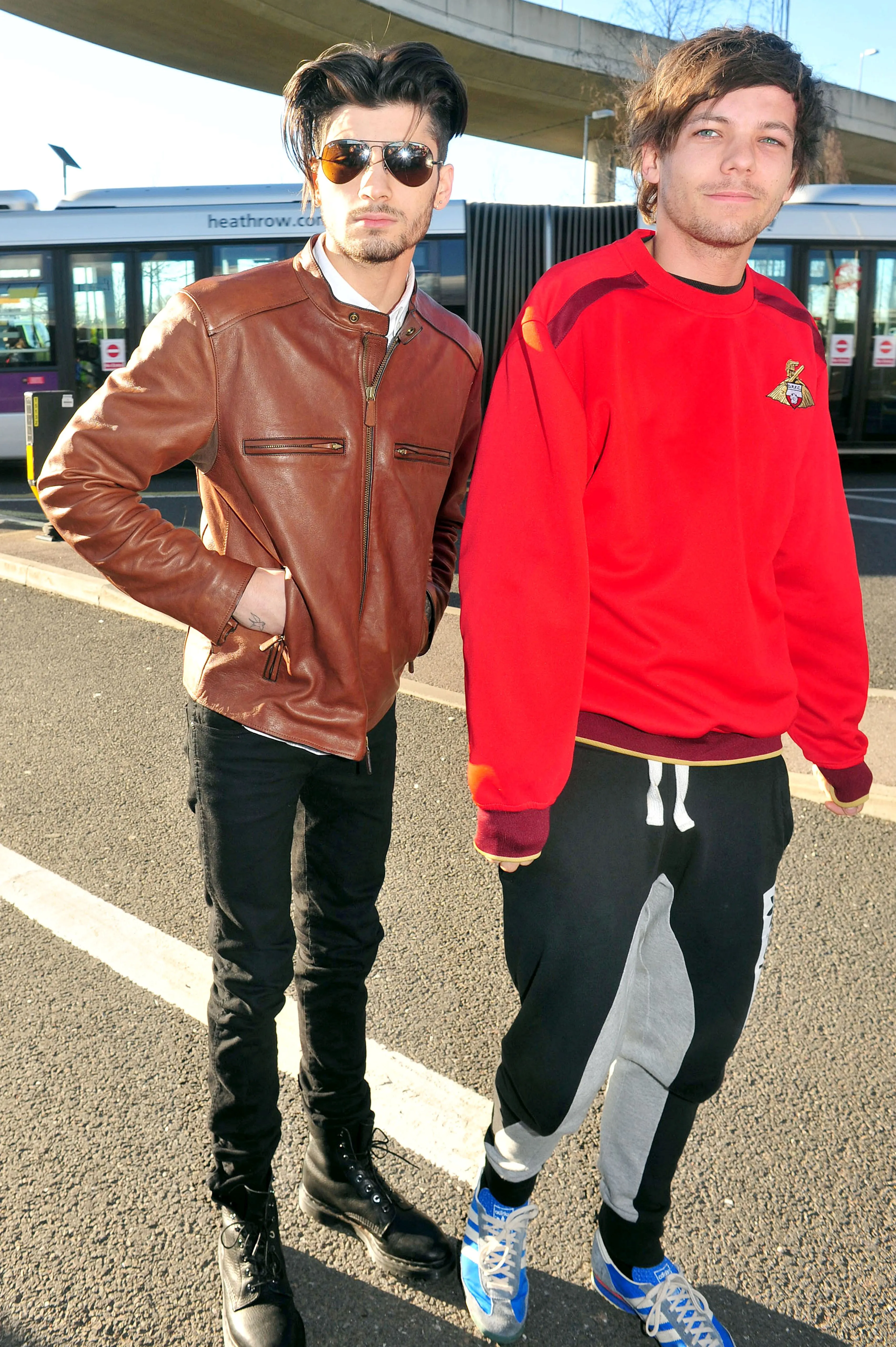 Zayn Malik in a brown jacket and Louis Tomlinson in a red shirt posing outdoors at Heathrow Airport.