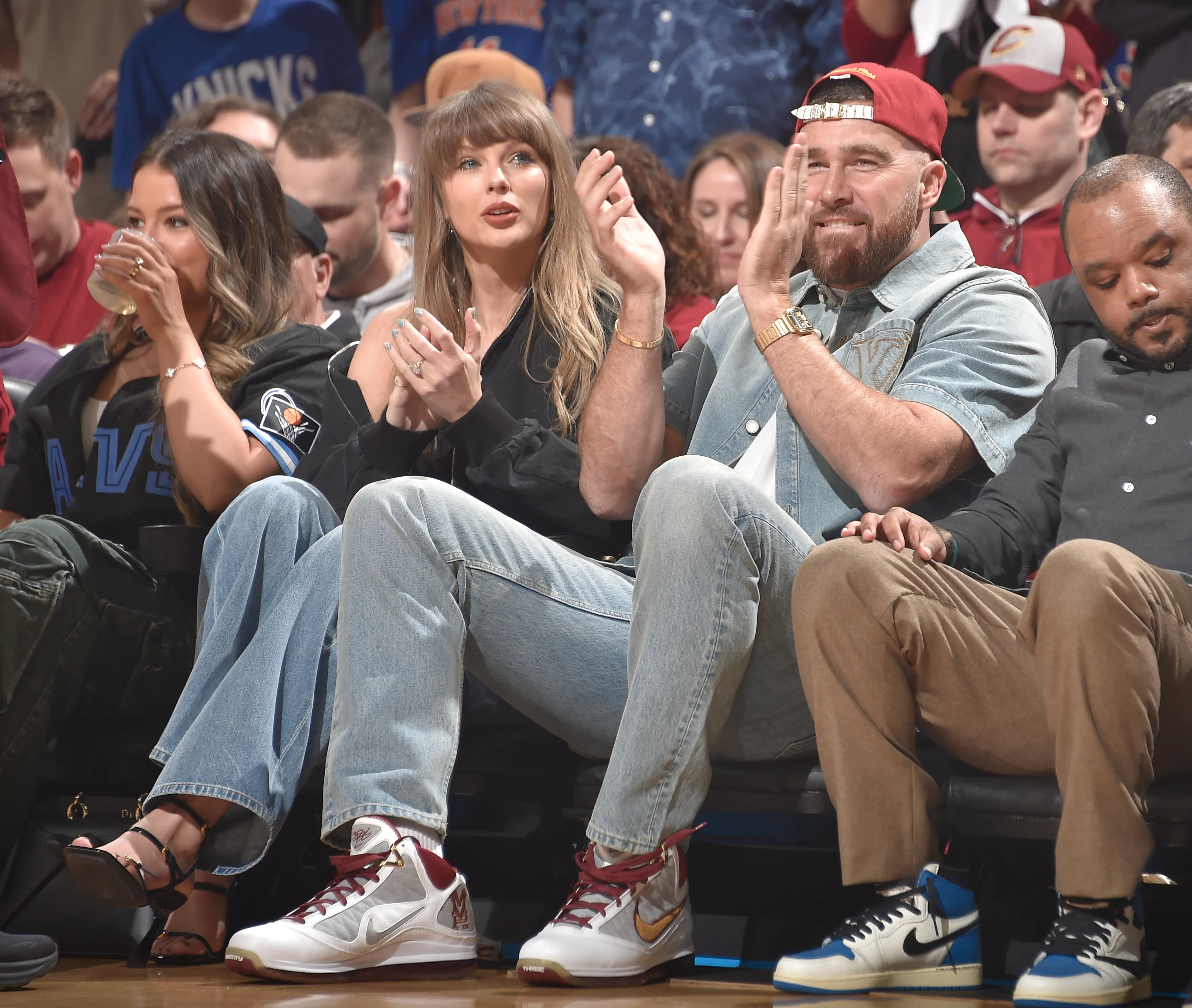 Taylor Swift and Travis Kelce clap while sitting courtside at a basketball game.