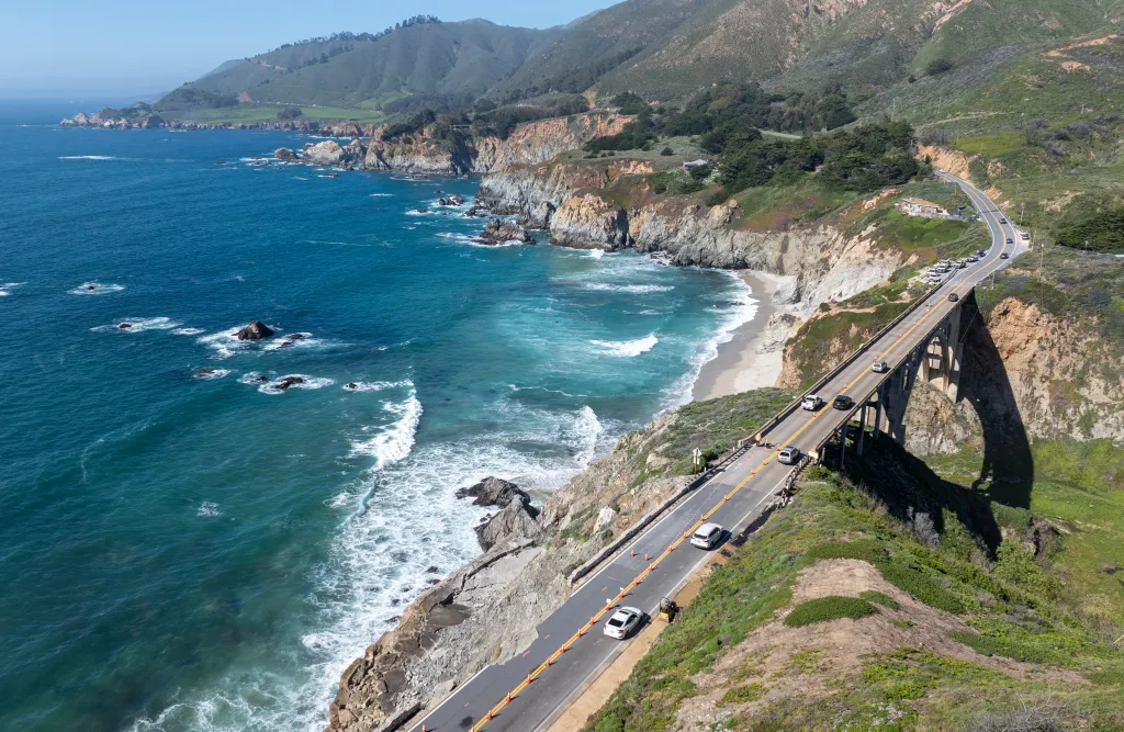 Aerial view of vehicles being escorted on a closed section of Highway 1 in Big Sur, California, next to the ocean.