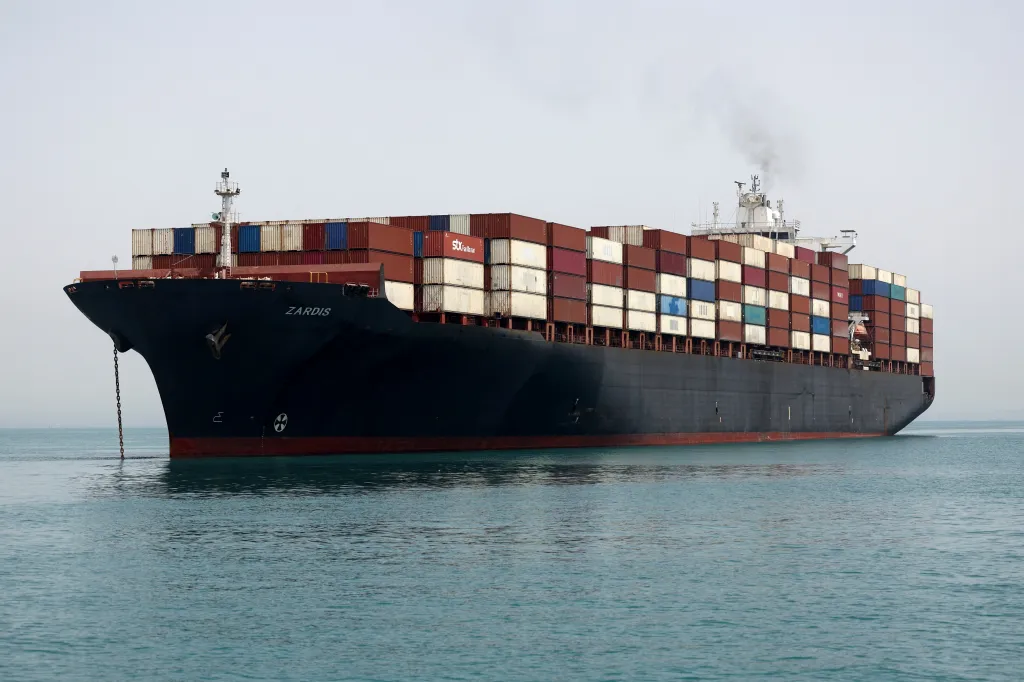 A cargo vessel, the ZARDIS, laden with shipping containers, moves through the Strait of Hormuz.