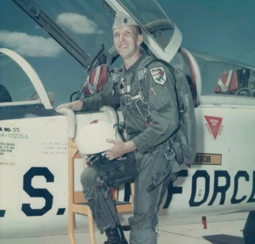 Ed Ashman, a veteran and teacher, wearing a flight suit and cap, smiling next to an Air Force jet.