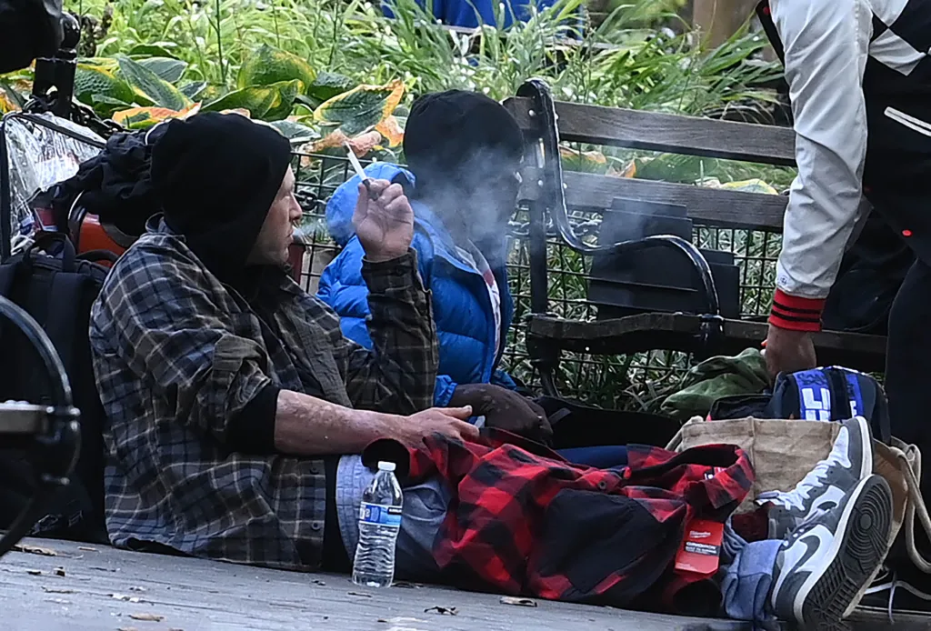 Two individuals smoking while sitting on the ground with belongings beside them.