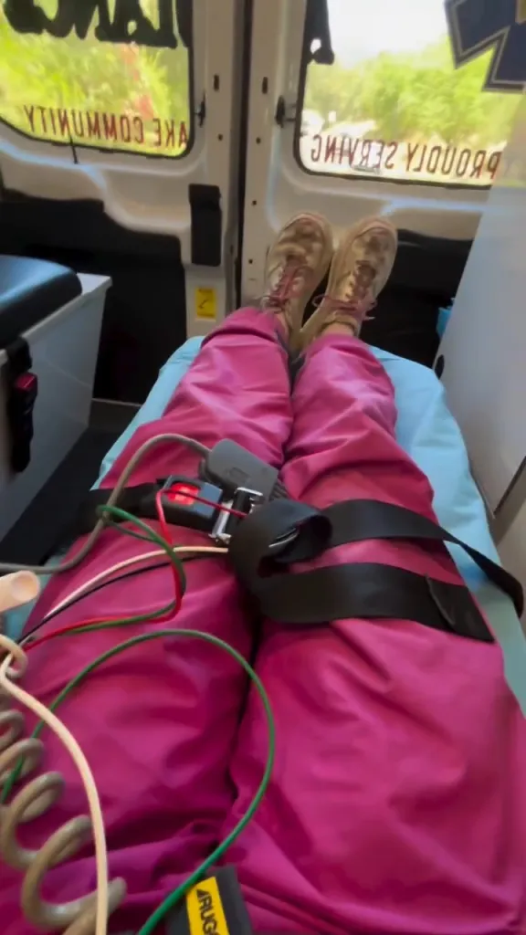 A person lying in an ambulance, wearing pink pants and hiking boots, with medical equipment on their legs.
