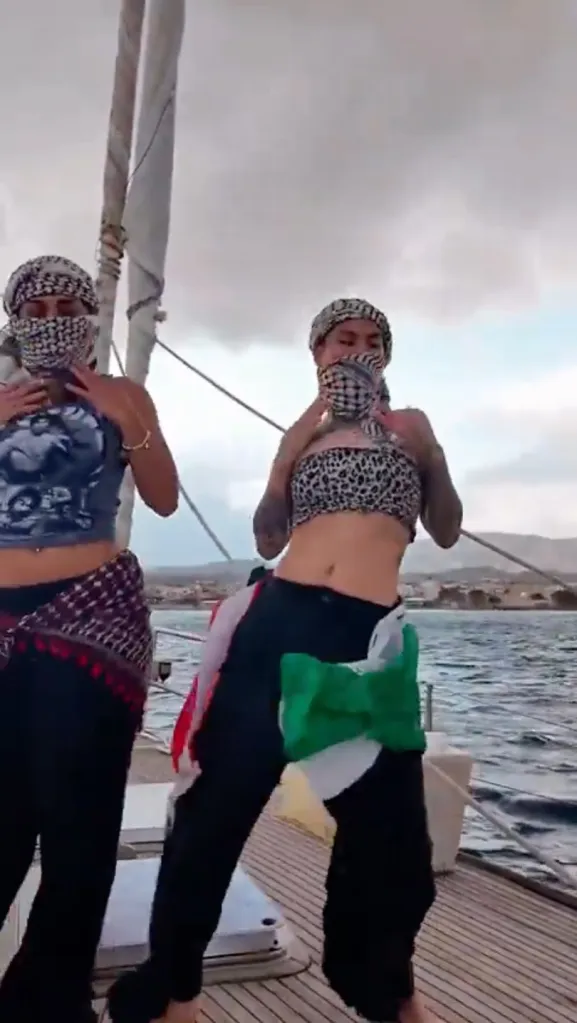 Two women dancing on a boat, wearing headscarves covering their faces, one with a Palestinian flag around her hips.