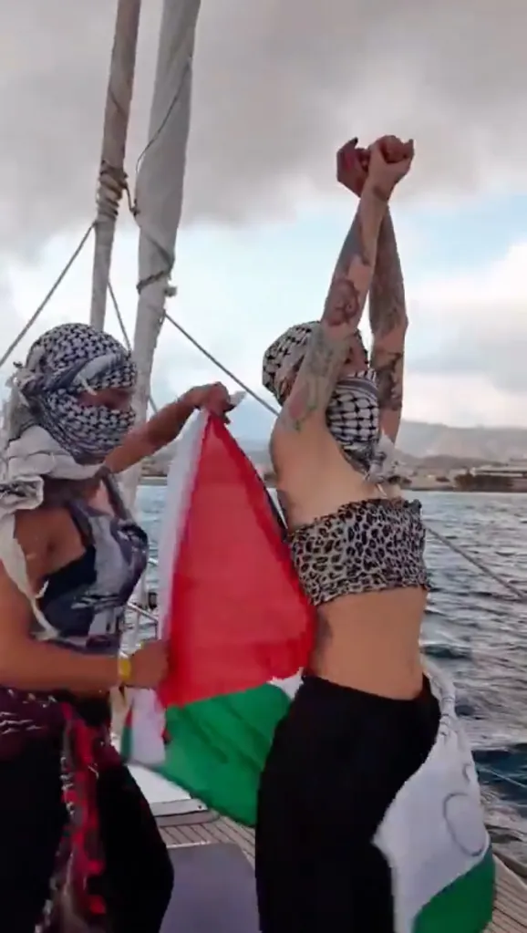 Two women on a boat, one holding a Palestinian flag and the other wearing one around her waist.