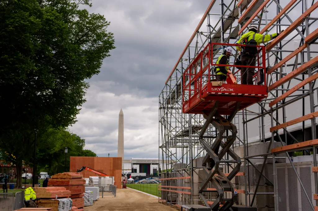 Two workers on a red scissor lift construct scaffolding on the National Mall with the Washington Monument in the background.