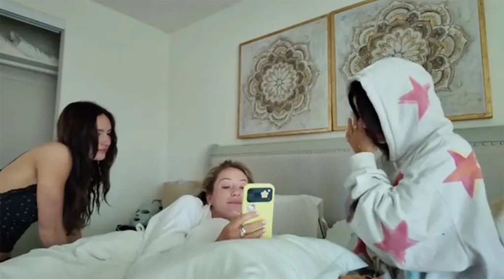 Alix Earle in bed looking at her phone, while a woman in a black top kneels beside her and another woman in a white star-print hoodie sits next to the bed.