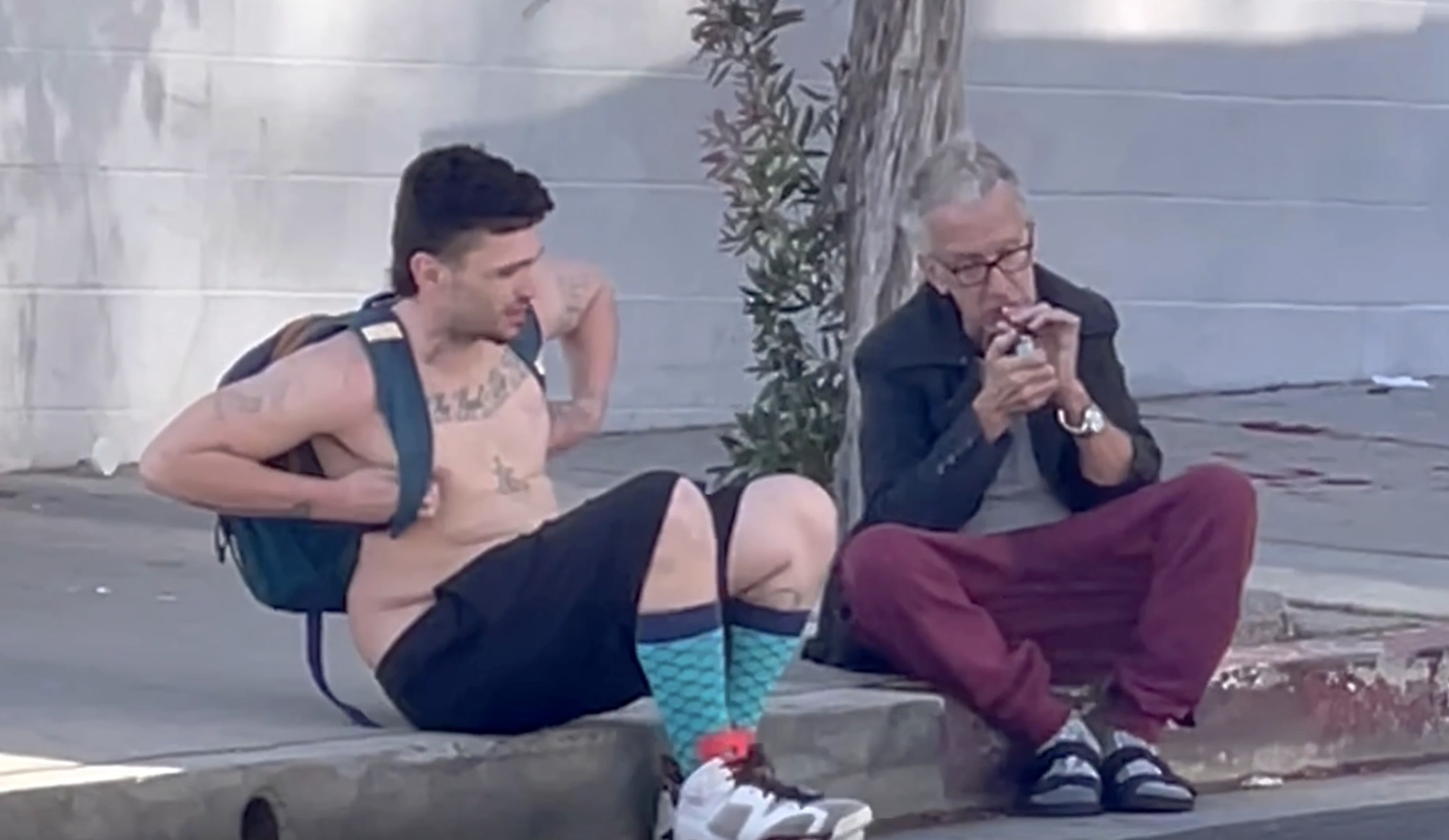 Andy Dick sitting on a curb next to another man, both looking disheveled, as Dick lights a cigarette.