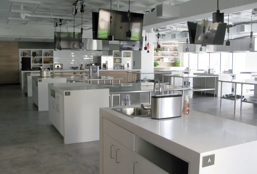 A bright, modern commercial kitchen with multiple cooking stations, each featuring a sink, stainless steel counters, and overhead screens.