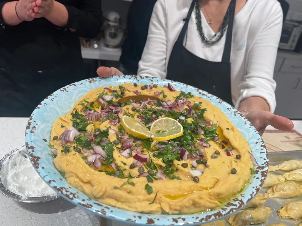 Person holding a platter of hummus garnished with red onions, capers, parsley, and lemon slices.