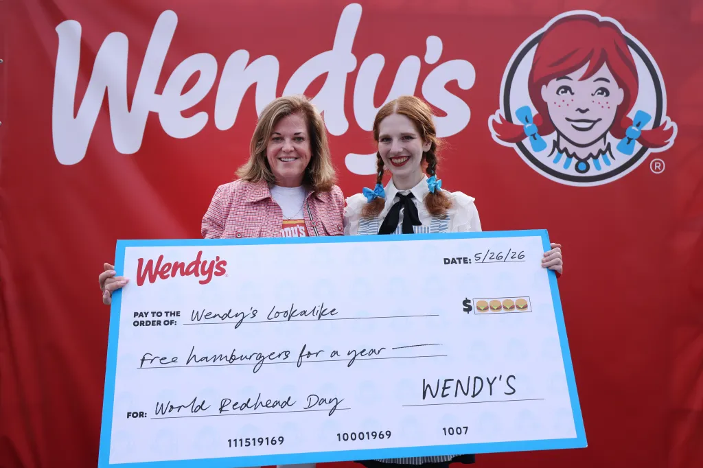 A woman in a red and white checkered shirt and a young woman dressed as Wendy from the Wendy's logo hold a large check for free hamburgers for a year.