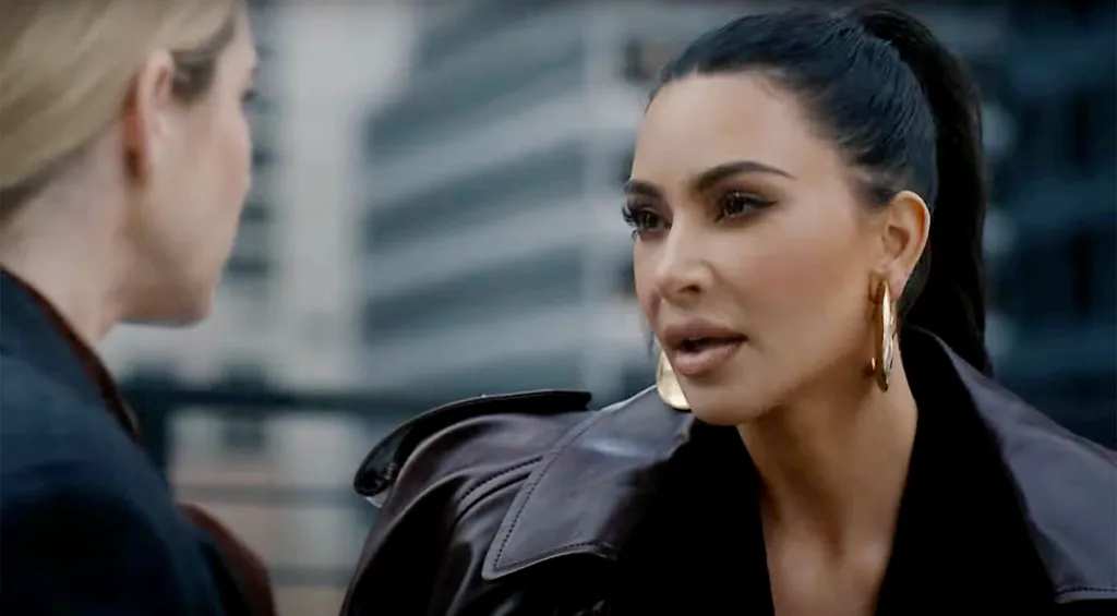 Kim Kardashian speaking while wearing a brown leather jacket and large gold hoop earrings.