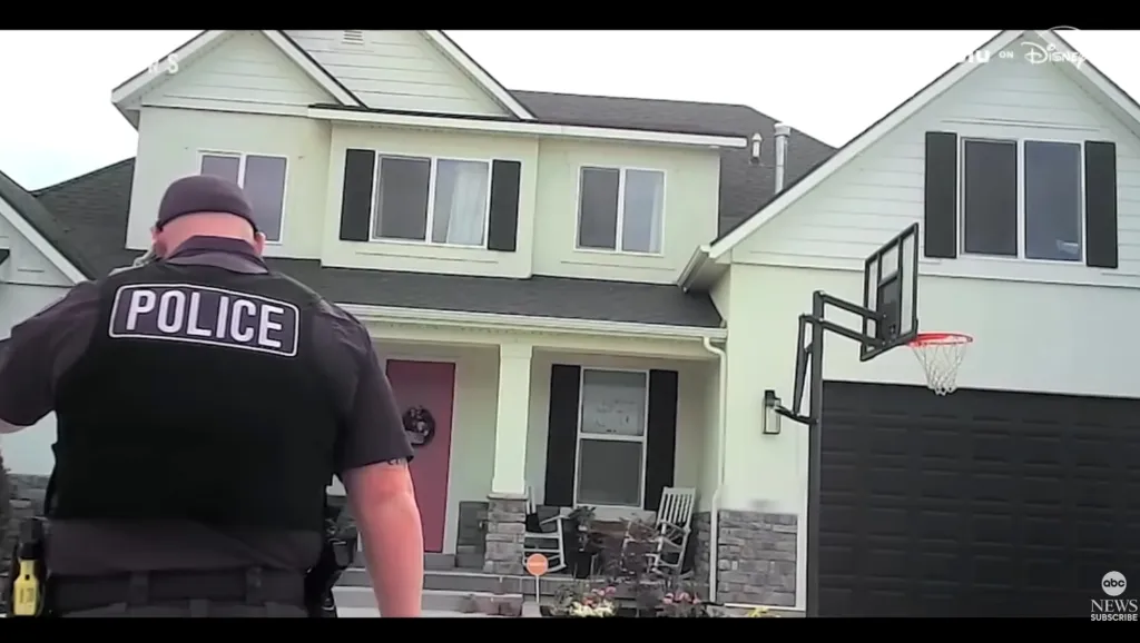 A police officer's back to the camera, facing a suburban house.