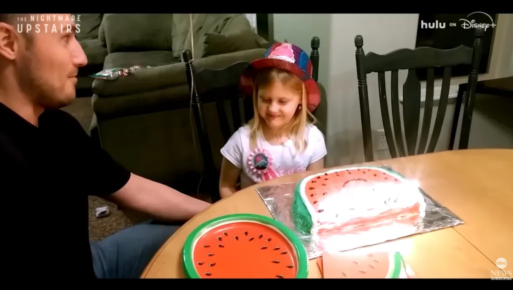 A man and a young girl at a table with a watermelon-shaped cake and plates.
