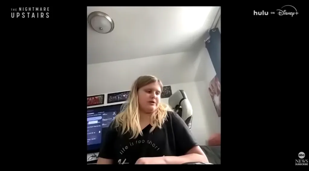 A blonde woman speaking to the camera in a room with a monitor and framed pictures.