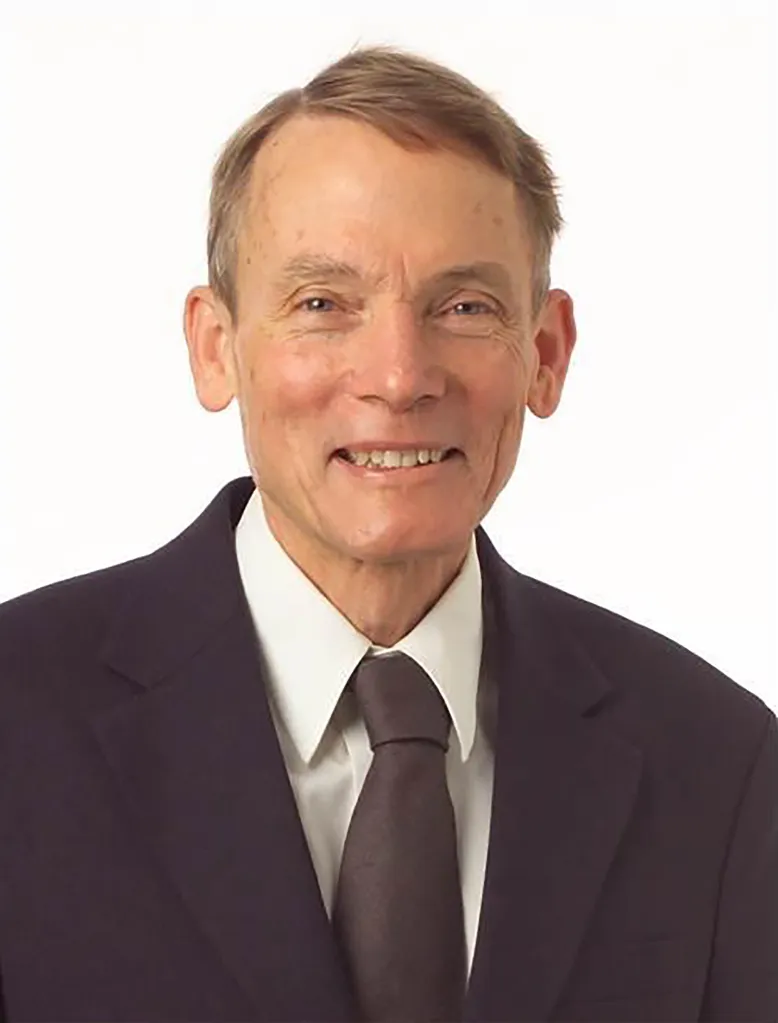 Headshot of William Happer, an older man with light brown hair, wearing a dark suit, white shirt, and dark tie, smiling softly against a white background.