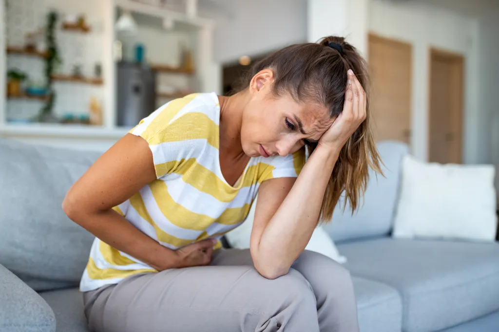 Woman sitting on a couch, holding her head and stomach in pain, suffering from migraine and abdominal cramps.